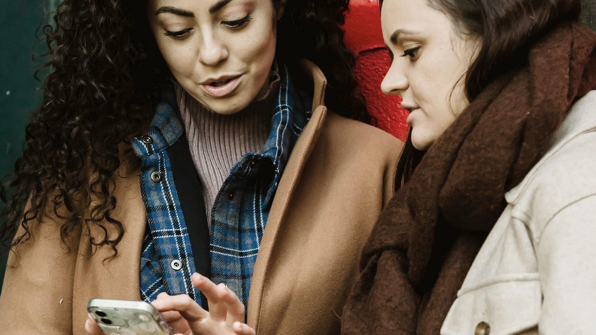 Två kvinnor i 30-40 årsåldern tittar på en mobil och diskuterar något. Dden ena kvinnan har mört lockigt hår och den andra har rakt mörkt hår