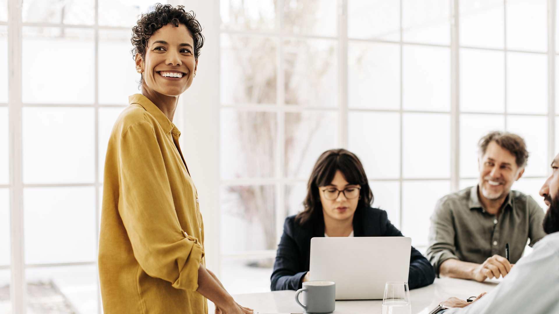 Människor sitter runt ett konferensbord. En kvinna står upp. Alla ler utom en kvinna sin skriver koncentrerat på sin dator.