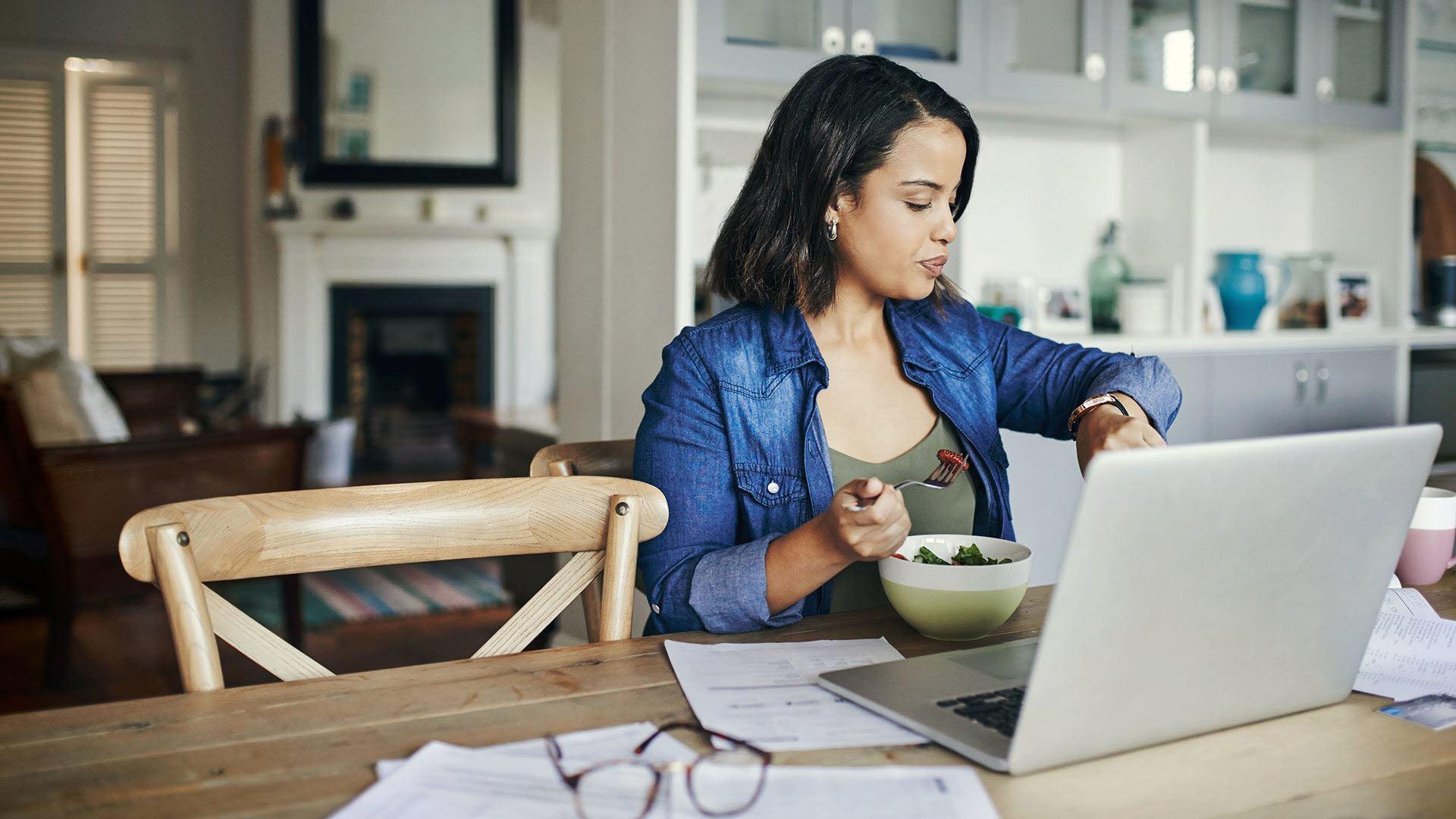 Latinsk mörkhårig kvinna 30-40 års åldern med jeansskjorta och grönt linne, äter en sallad framför datorn samtidigt som hon tittar på klockan